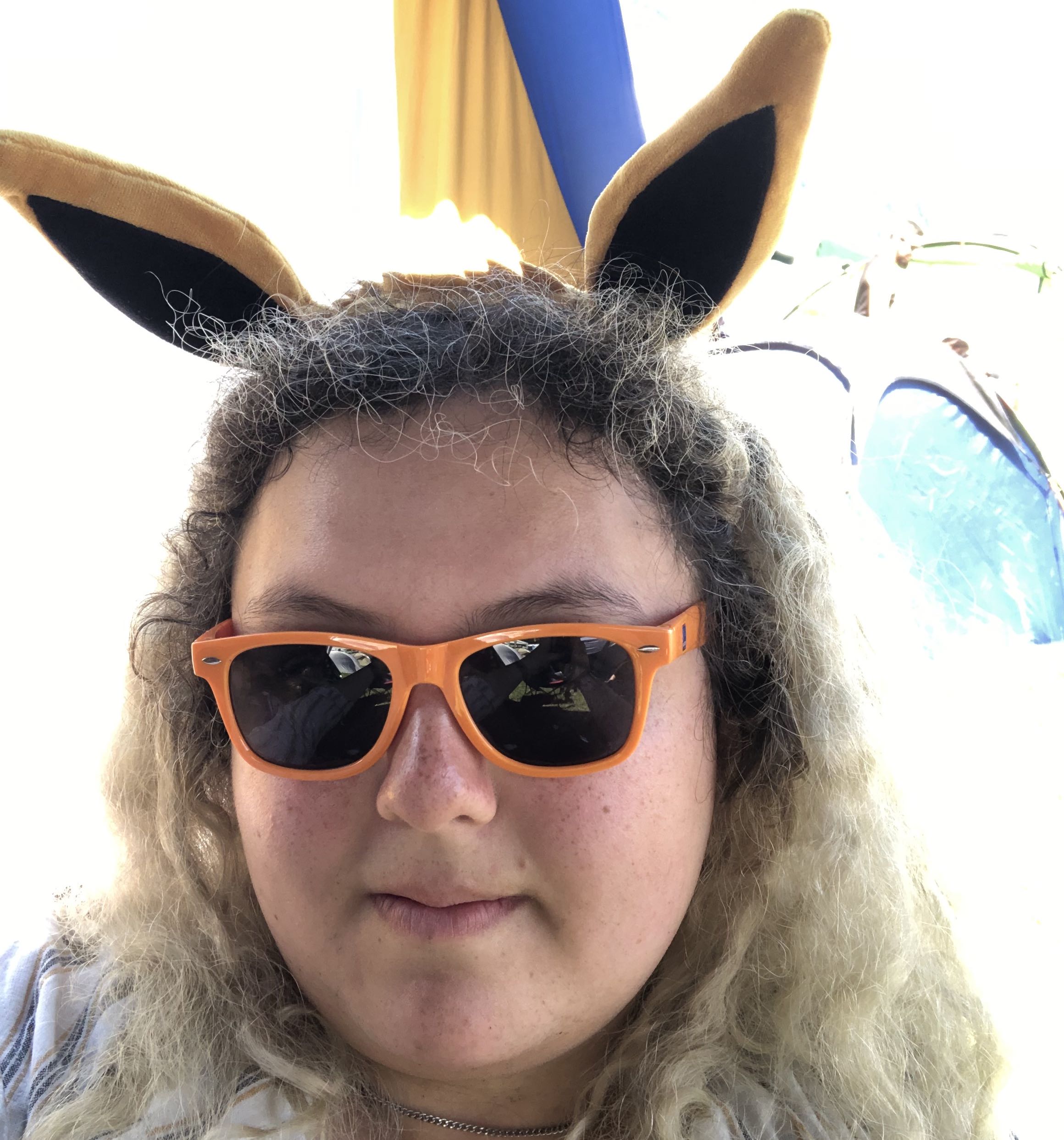 A head shot of a woman with messy hair, eevee ears, and orange sunglasses.